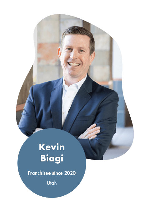 Kevin Biagi - Logistics Franchise Opportunity Reviews and Testimonials Smiling man in a blue suit with arms crossed, standing indoors. Text on the image reads Kevin Biagi, Franchisee since 2020, Utah. Background features a blurred building interior, embodying the success often highlighted in franchise reviews by owners.