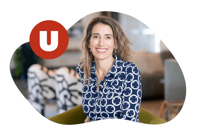 Franchise_Home_5050_4 A woman with wavy hair sits smiling, wearing a blue and white patterned blouse. A red circle with a white U is in the top left. The background shows a blurred room with patterned chairs and a couch.