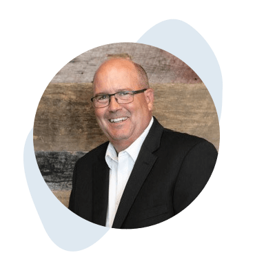 Franchise_Leadership_Tile_Bart A man wearing a black suit and white shirt is smiling. He has glasses and short hair. The background is a textured wall with wood-like colors.