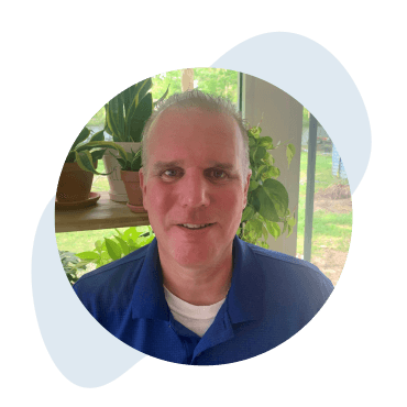 Franchise_Leadership_Tile_Chris A man in a blue shirt is smiling, with a backdrop of indoor plants and a window showing a glimpse of greenery outside.