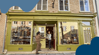 A delivery person from a shipping franchise hands a package to someone at the entrance of a shop with a green facade and large windows displaying various items. The store, part of the franchise, is set in a brick building with reflections of surrounding structures.