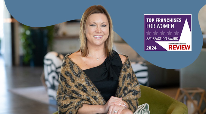 A woman smiling and sitting in an indoor setting. She is wearing a leopard print shawl and a black top. Next to her, theres a sign that reads Top Franchises for Women Satisfaction Award 2024 from Franchise Business Review.