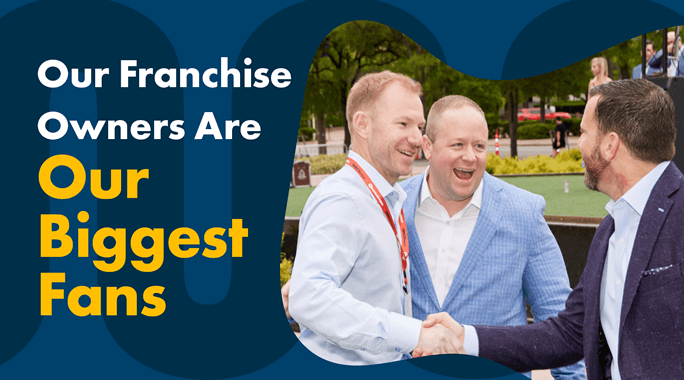Three men in suits are smiling and shaking hands outdoors at an event. The text reads, Our Franchise Owners Are Our Biggest Fans in bold white and yellow letters. Trees and people in the background suggest a park setting.