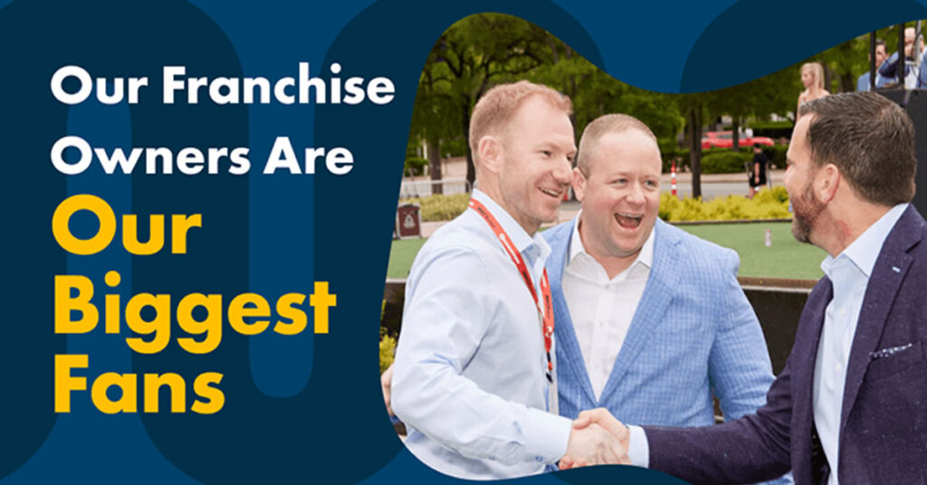 Three men in suits are smiling and shaking hands outdoors at an event. The text reads, Our Franchise Owners Are Our Biggest Fans in bold white and yellow letters. Trees and people in the background suggest a park setting.