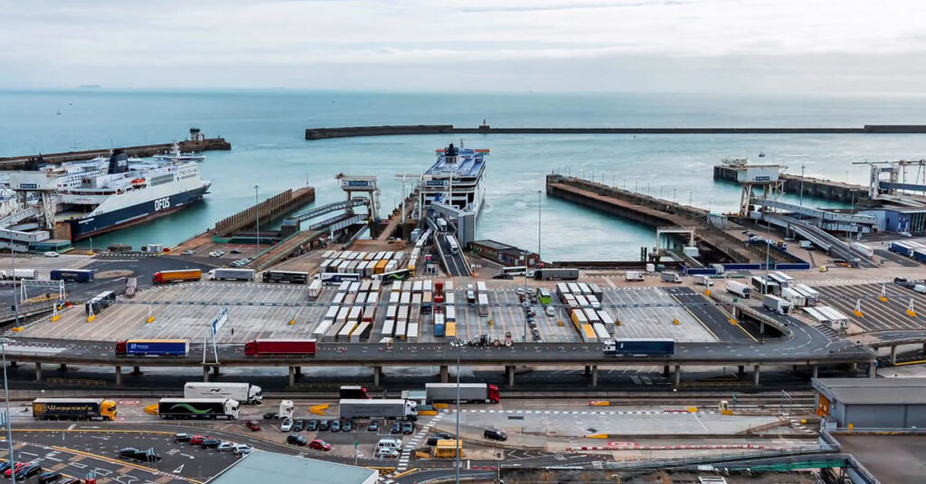 A busy ferry port with several ferries docked, rows of trucks and vehicles lined up, and the sea in the background under a cloudy sky. A red play button is overlaid at the center of the image.