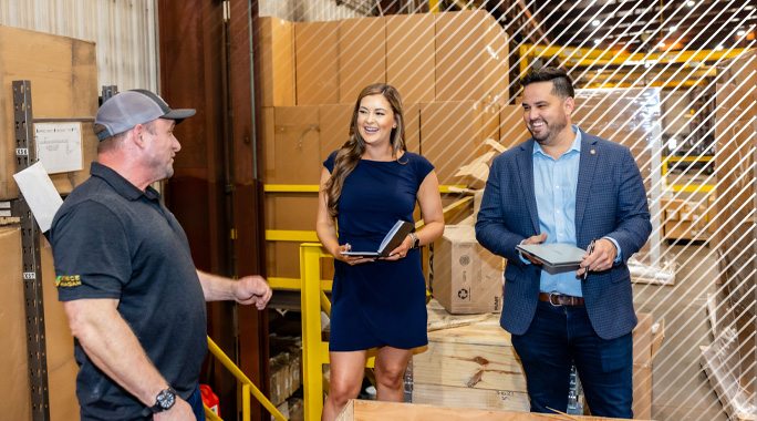 Three people stand in a warehouse, smiling and talking. One person wears a polo shirt and cap, while the other two, holding tablets, are dressed in business attire. Large cardboard boxes and equipment are visible in the background.