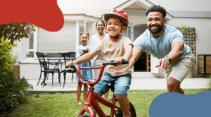 A smiling child wearing a helmet rides a red bicycle on the grass, assisted by a man guiding him. A woman and another child stand in the background near a house, joyfully watching. The scene conveys a cheerful family moment outdoors.