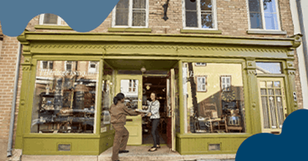 A delivery person from a shipping franchise hands a package to someone at the entrance of a shop with a green facade and large windows displaying various items. The store, part of the franchise, is set in a brick building with reflections of surrounding structures.