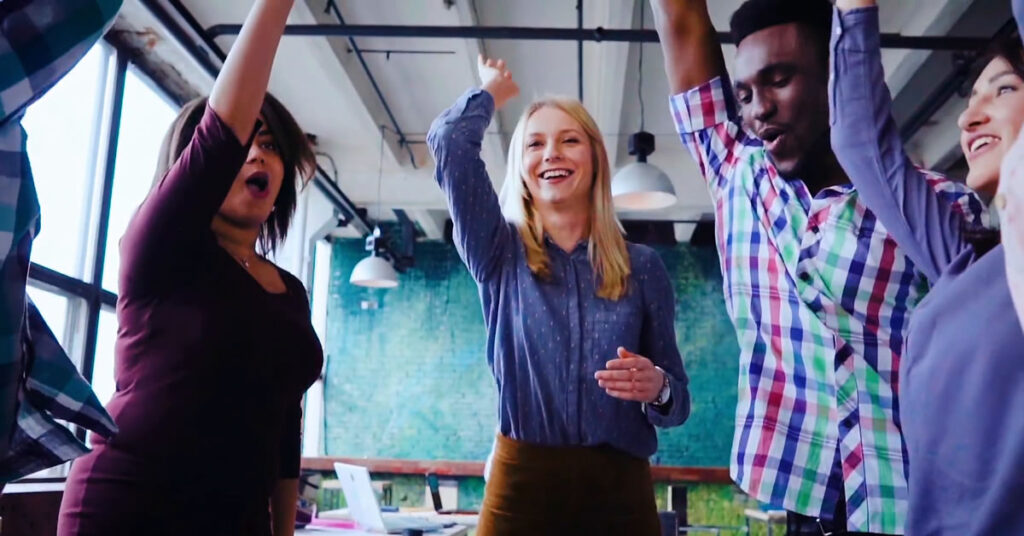 A diverse group of four people stand in an office, smiling and raising their arms in celebration. A red play button is centered over the image, suggesting it is a video thumbnail.