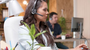 A woman wearing a headset sits at a desk, focused on her computer, epitomizing SMB success in a bustling office. Nearby, a man also dons a headset while working diligently. Around them, plants and office equipment add life to their Unishippers workspace.