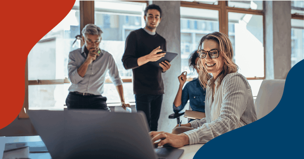 Four people in a bright office, three standing and talking in the background while one woman sits at a desk in the foreground, smiling at the camera and working on a laptop.