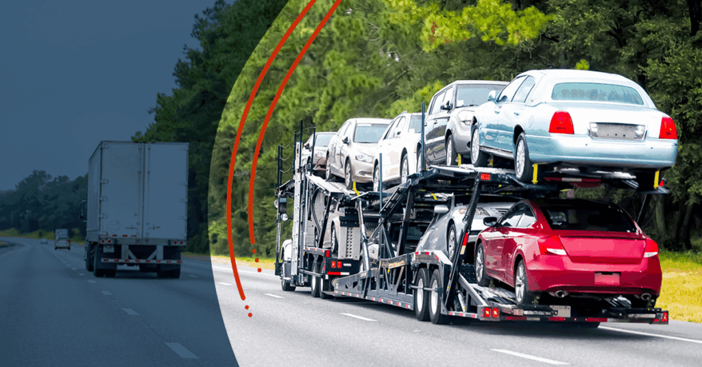 A car carrier truck transports multiple vehicles on a highway, alongside a semi-truck, with trees and greenery lining the road.