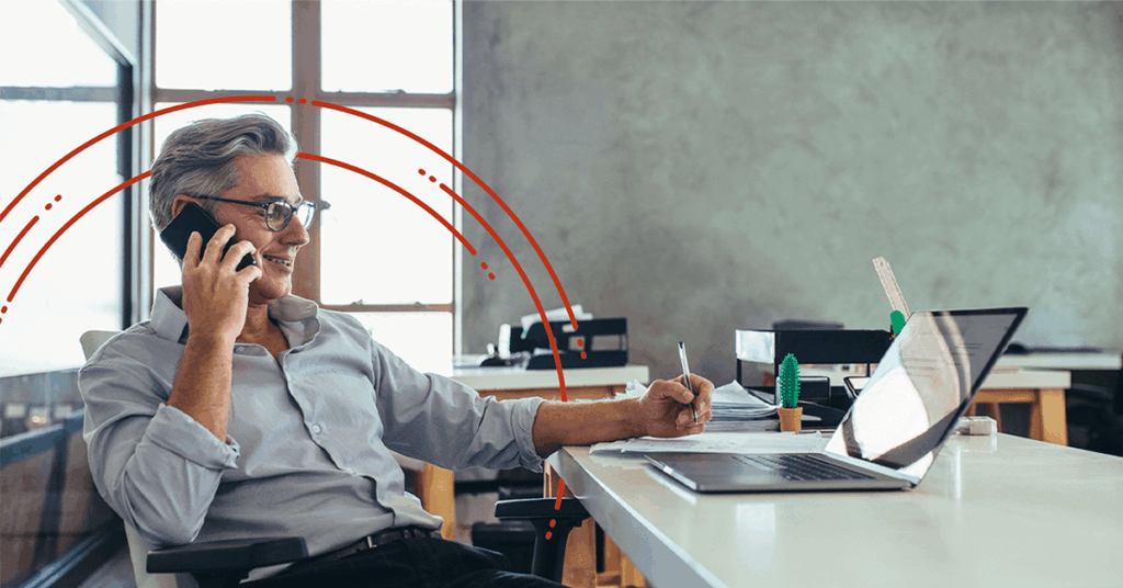 A smiling man with gray hair sits at a desk in an office, talking on the phone, taking notes, and working on a laptop. Red graphic lines arch around him, and sunlight streams through a window behind him.