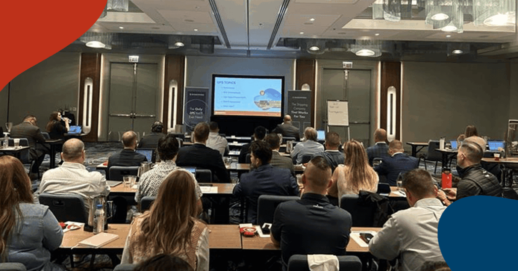 A group of people sit at tables facing a projector screen in a modern conference room, attending a presentation. Most attendees are focused on the speaker and presentation slides at the front.