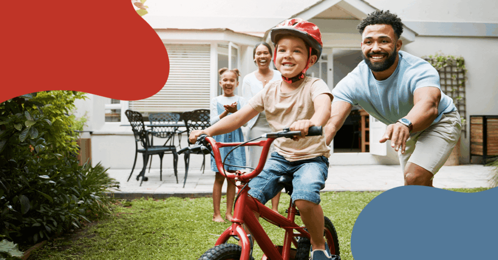 A smiling child wearing a helmet rides a red bicycle on grass while a man helps guide them. Two people stand behind, smiling, outside a house with a patio table and chairs.
