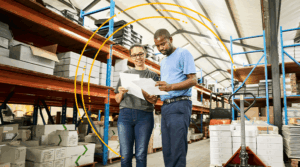 Two people stand in a warehouse, reviewing documents together. Shelves filled with boxes and supplies surround them, and yellow graphics form a partial circle around the pair, highlighting their collaboration.