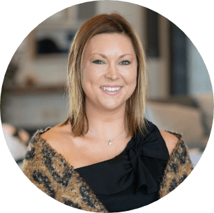 A woman with straight, shoulder-length blonde hair smiles at the camera. She is wearing a black, one-shoulder top with a bow and a brown patterned shawl, with a softly blurred indoor background.