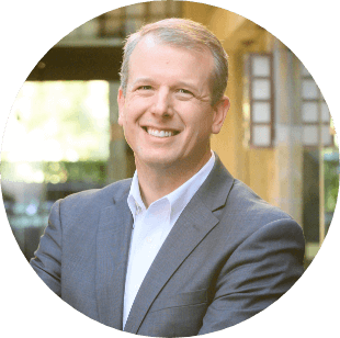 A man in a suit jacket and dress shirt smiles while standing outdoors with blurred buildings and greenery in the background.