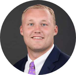 A smiling man with short blond hair wearing a dark suit, white shirt, and purple patterned tie, posed in front of a plain dark background.
