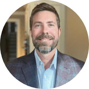 A man with short brown hair and a beard, wearing a blue checked blazer and light blue shirt, smiles while standing indoors in a well-lit room with a blurred background.