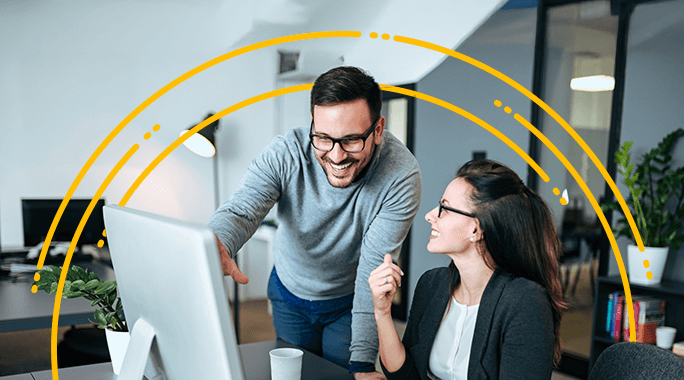 Two colleagues in an office smile and discuss something on a computer screen related to MB Shipping Customers. One stands and points at the monitor while the other sits holding a pen, with yellow arcs digitally overlaid above them.