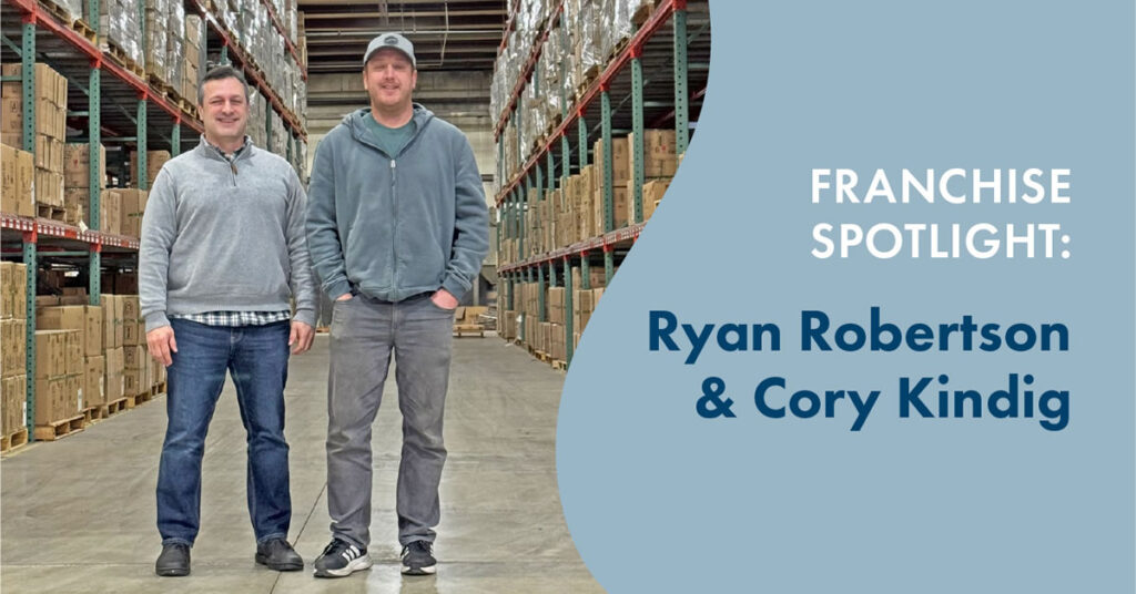 Two men stand smiling in a large warehouse filled with shelves of boxes. Text on the right reads, Unishippers Franchise Spotlight: Ryan Robertson & Cory Kindig on a blue background.