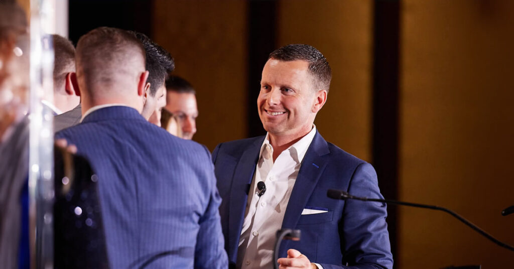 A man in a blue suit, smiling and holding a microphone, talks with a group of people in formal attire at an indoor event or conference.