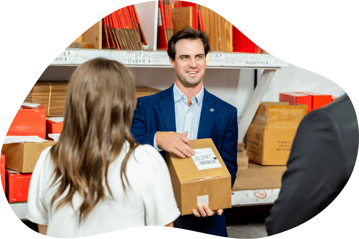 A man in a blue suit hands a package to a woman with long brown hair in a warehouse filled with shelves of boxes and red folders, reflecting the dynamic 3PL growth in the logistics industry.