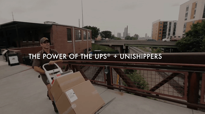 A delivery person pushes a hand truck loaded with boxes along an outdoor walkway, with city buildings and a bridge in the background. Text overlay reads: THE POWER OF THE UPS® + UNISHIPPERS.