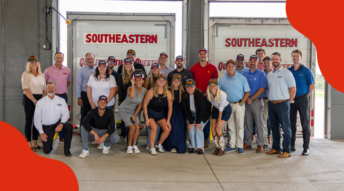 A group of people posing and smiling in front of two Southeastern Freight Lines trucks inside a warehouse, wearing business casual attire and hats—highlighting the team spirit behind successful shipping franchise opportunities.