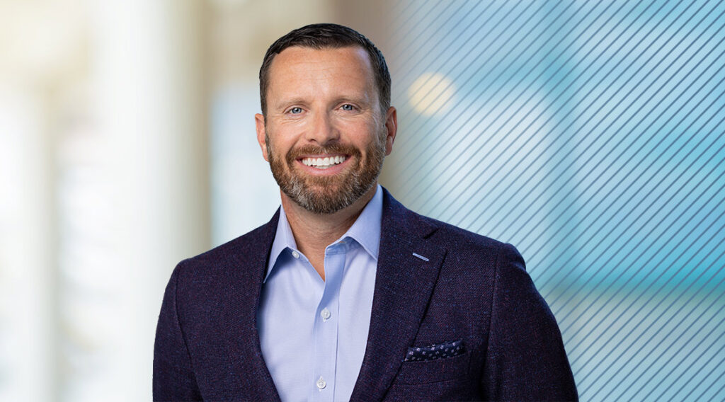 Smiling man with short brown hair and a beard, wearing a light blue dress shirt and dark blazer, stands in front of a blurred background with diagonal lines on the right side, reflecting the dynamic world of logistics technology.